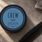 American Crew Fiber 50 g z niebieską etykietą na kamiennym blacie obok czarnego grzebienia.