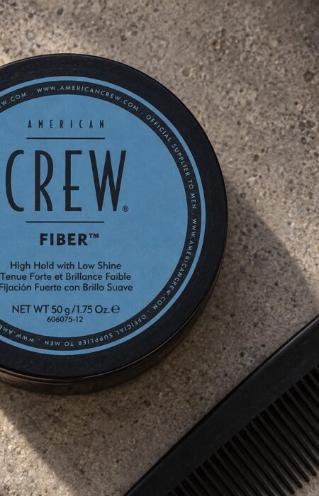 American Crew Fiber 50 g z niebieską etykietą na kamiennym blacie obok czarnego grzebienia.