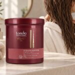 Bordowy słoik Londa Velvet Oil In-Salon Treatment obok ręcznika, w tle kobieta z wilgotnymi włosami