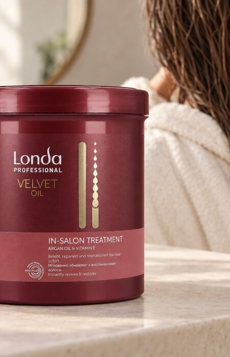 Bordowy słoik Londa Velvet Oil In-Salon Treatment obok ręcznika, w tle kobieta z wilgotnymi włosami