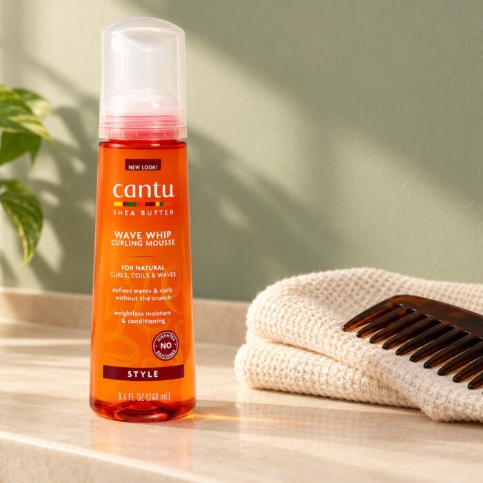 Pomarańczowa butelka Cantu Wave Whip Curling Mousse 248 ml obok ręcznika i grzebienia
